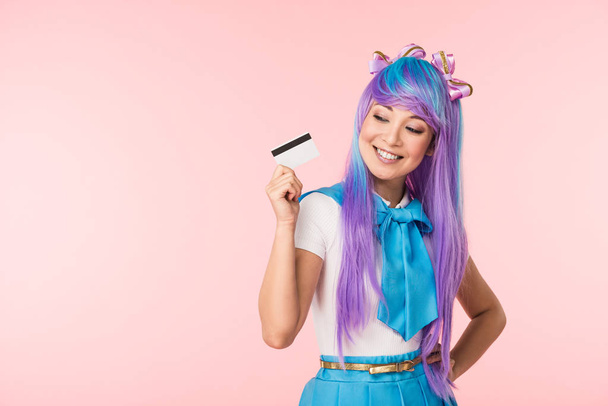 Smiling asian anime girl in purple wig holding credit card isolated on pink - Zdjęcie, obraz