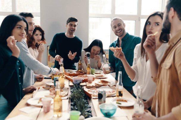 Group of young people in casual wear eating pizza and smiling while having a dinner party indoors - 写真・画像