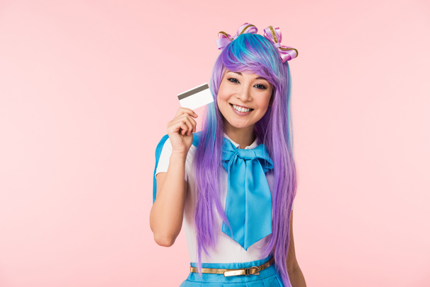 Glimlachend Aziatische anime meisje in paarse pruik Holding creditcard geïsoleerd op roze - Foto, afbeelding