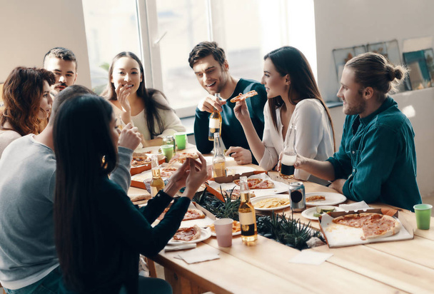 Group of young people in casual wear eating pizza and smiling while having a dinner party indoors - Foto, Imagem