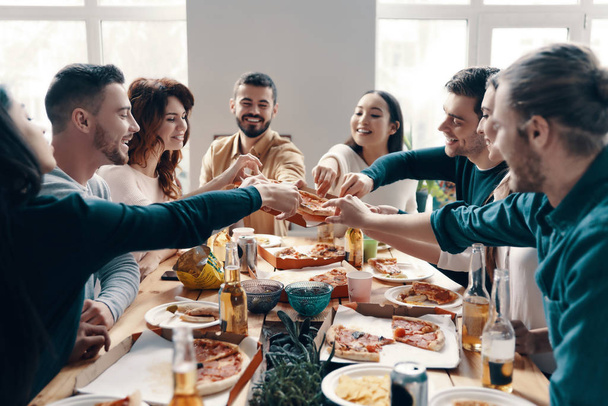 Group of young people in casual wear picking pizza and smiling while having a dinner party indoors - 写真・画像