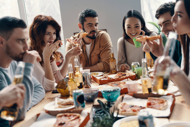 Happy to be around. Group of young people in casual wear eating pizza and smiling while having a dinner party indoors  - Photo, Image