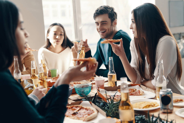 Great food and great people. Group of young people in casual wear eating pizza and smiling while having a dinner party indoors - Фото, изображение