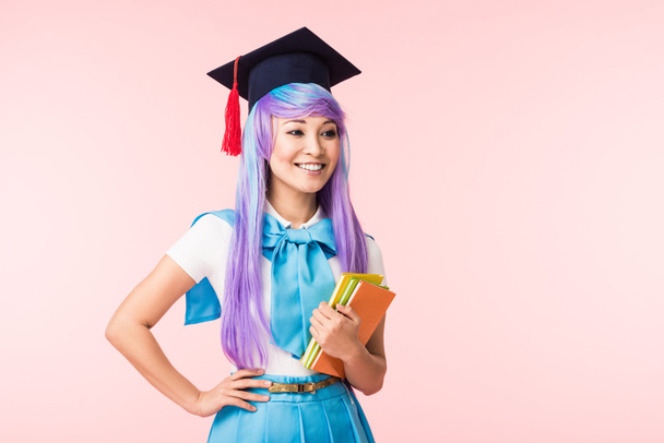 Smiling asian anime girl in academic cap holding books isolated on pink - Photo, Image