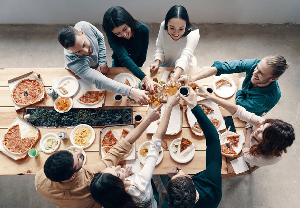 Top view of young people in casual wear toasting each other and smiling while having a dinner party indoors - Фото, зображення