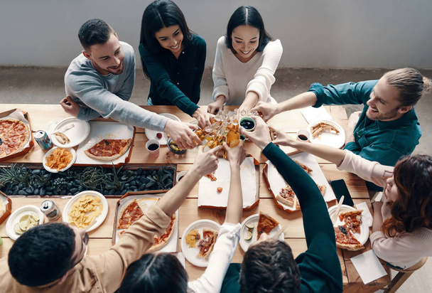 Top view of young people in casual wear toasting each other and smiling while having a dinner party indoors    - Foto, imagen