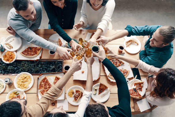 Top view of young people in casual wear toasting each other and smiling while having a dinner party indoors             - Foto, immagini