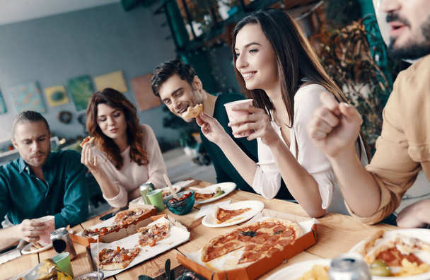 glücklich, dabei zu sein. Gruppe junger Leute in Freizeitkleidung plaudert und lächelt bei einer Dinnerparty drinnen  - Foto, Bild