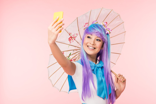 Χαμογελαστός Ασιάτης anime κορίτσι με χαρτί ομπρέλα παίρνοντας selfie απομονωμένη σε ροζ - Φωτογραφία, εικόνα