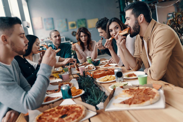 Hyvää ruokaa ja seuraa. Ryhmä nuoria rennoissa vaatteissa syömässä pizzaa ja hymyilemässä illalliskutsuilla sisätiloissa
 - Valokuva, kuva