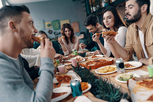 Хорошие времена с друзьями. Группа молодых людей в повседневной одежде едят пиццу и улыбаются во время ужина в помещении
 - Фото, изображение