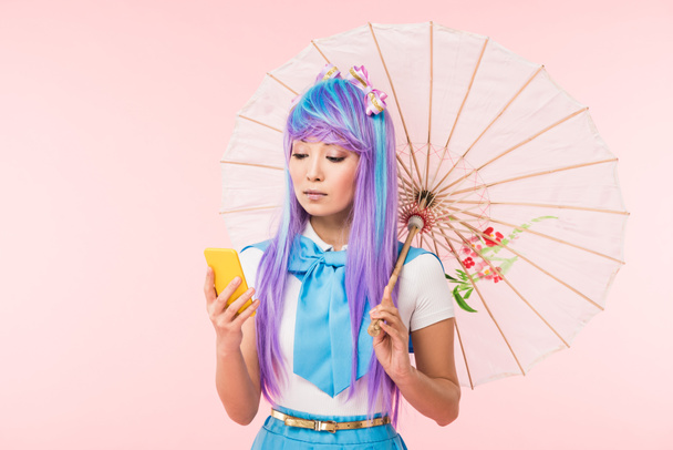 Ασιάτης/ισσα anime κορίτσι κρατώντας χαρτί ομπρέλα και χρησιμοποιώντας smartphone απομονωθεί σε ροζ - Φωτογραφία, εικόνα