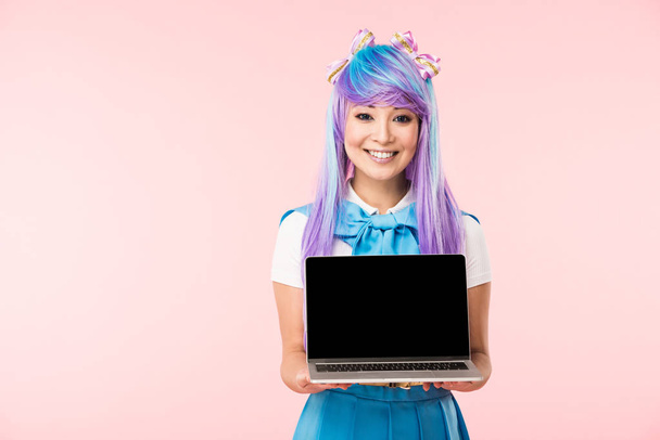 Lachende Aziatische anime meisje in pruik toont laptop met blanco scherm geïsoleerd op roze - Foto, afbeelding