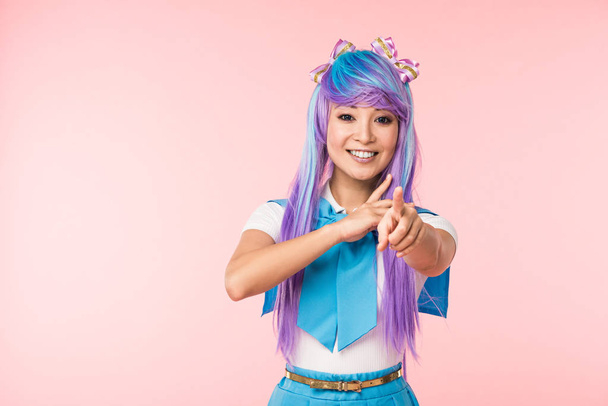 Smiling asian anime girl in purple wig pointing with finger at camera isolated on pink - 写真・画像
