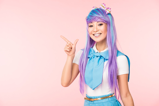 Glimlachend Aziatische anime meisje in paarse pruik wijzend met vinger geïsoleerd op roze - Foto, afbeelding