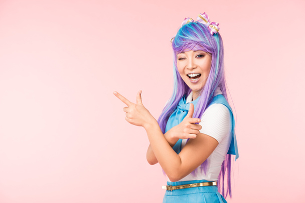 Joyful anime girl in purple wig pointing with fingers and winking isolated on pink - Foto, imagen