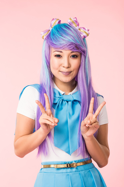 Smiling anime girl in purple wig showing peace signs isolated on pink - Photo, image