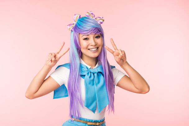Radostná Asijská anime dívka v purpurové paruce ukazující mírové znaky izolované na růžovém - Fotografie, Obrázek