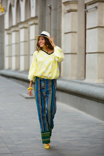Brunette girl dressed in yellow in white peas blues, striped pants and hat walks in the city street on a sunny day - Foto, Imagen
