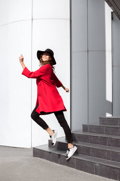 Charming brunette girl dressed in bright red coat and fashionable hat with wide brim poses in the street - Фото, изображение