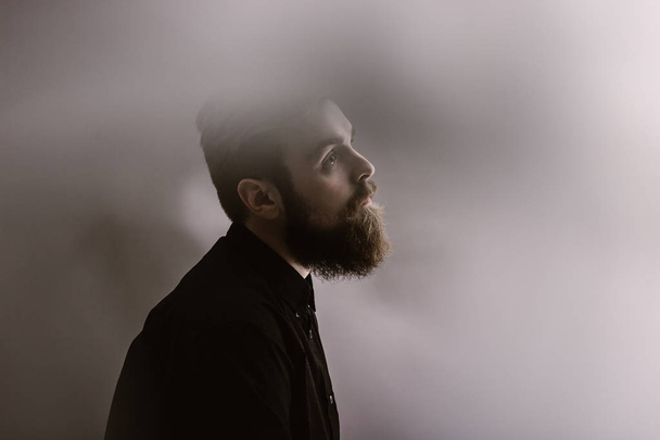 Sepia photo portrait of a stylish man in profile with a beard dressed in the black shirt in the fog - 写真・画像