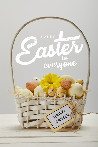 wicker basket with colorful painted eggs, yellow gerbera flower, greeting card and happy Easter to everyone lettering on grey background - Photo, Image