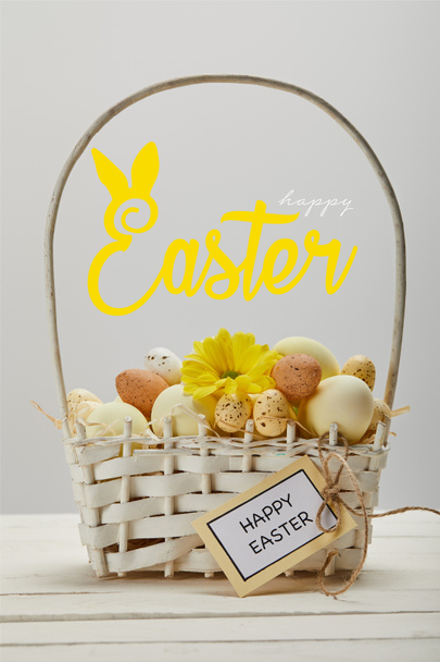 wicker basket with colorful painted eggs, yellow gerbera flower, greeting card and happy Easter lettering on grey background - Photo, Image