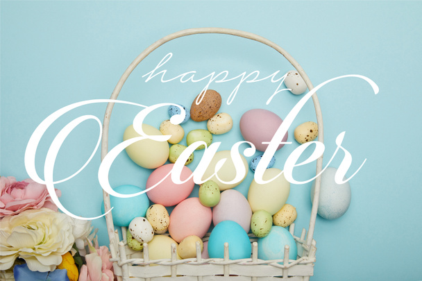 top view of painted multicolored eggs scattered near wicker basket and flowers on blue background with happy Easter lettering - Foto, Imagen