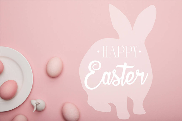 top view of painted eggs and white decorative bunny near plate on pink background with happy Easter lettering - Fotó, kép