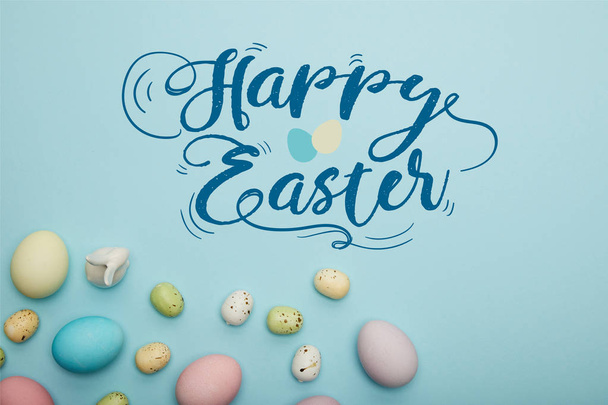 top view of painted multicolored eggs scattered near decorative rabbit on blue background with happy Easter lettering - Photo, image