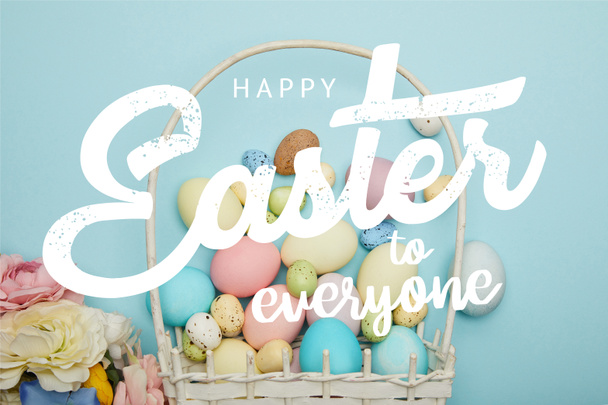 top view of painted multicolored eggs, wicker basket and flowers on blue background with happy Easter to everyone lettering - Photo, Image