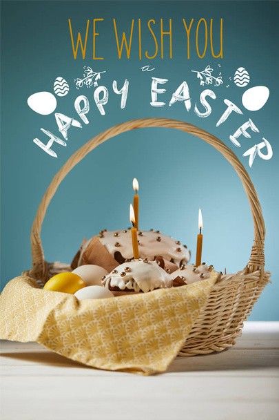 traditional Easter cakes with burning candles and chicken eggs in wicker basket on blue background with we wish you happy easter lettering - 写真・画像