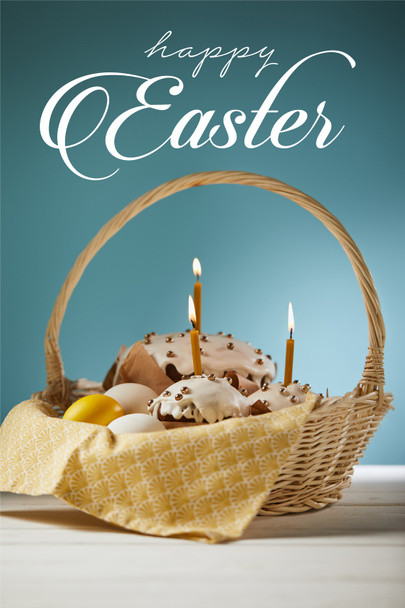traditional Easter cakes with burning candles and chicken eggs in wicker basket on blue background with happy easter lettering - 写真・画像
