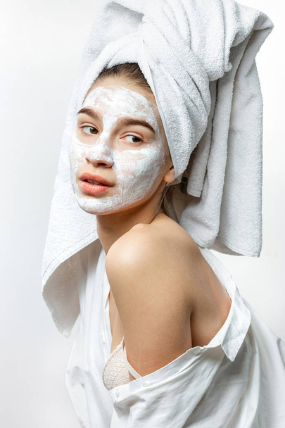 Beautiful young woman dressed in white clothes with a white towel on her hair and cosmetic mask on her face holds her hand on her naked shoulder - Foto, immagini