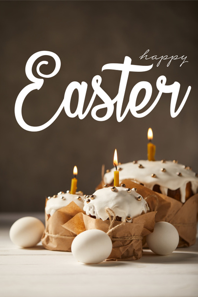 traditional tasty Easter cakes with burning candles and white chicken eggs on brown background with happy easter lettering - Photo, Image
