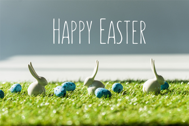 decorative bunnies on green grass near blue quail eggs with happy Easter lettering - Foto, afbeelding