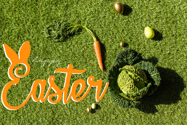 top view of carrot, savoy cabbage and painted Easter eggs on green grass with happy Easter lettering - Photo, Image
