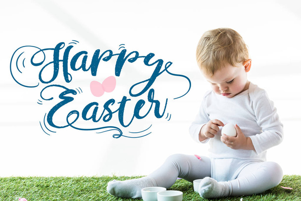 child sitting with white chicken egg on green grass near blue happy Easter lettering - Fotoğraf, Görsel