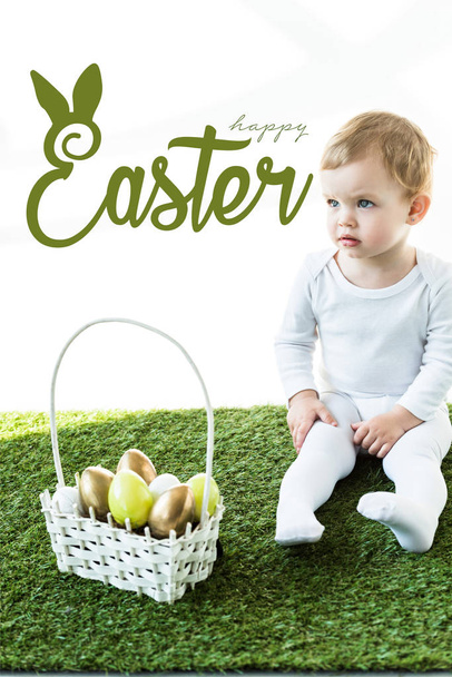 child sitting on green grass near basket with Easter eggs and happy Easter lettering above - Foto, Bild