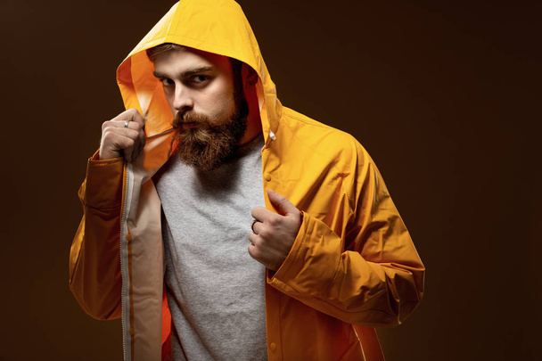 Red-haired guy with a beard and mustache dressed in a gray t-shirt and yellow jacket with a hood is posing on a brown background in the studio - Photo, image