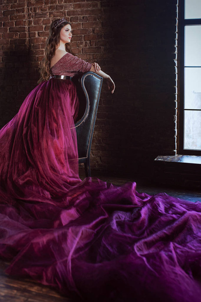 πορτρέτο του όμορφο νεαρό όμορφο χαριτωμένο γυναίκα πριγκίπισσα (βασίλισσα) στο μακρύ μωβ φόρεμα της Βασίλισσας και στέμμα, με μακριά μαλλιά και να κάνει εσωτερικό σε μινιμαλιστικό εσωτερικό σοφίτα στέκεται δίπλα στο παράθυρο - Φωτογραφία, εικόνα