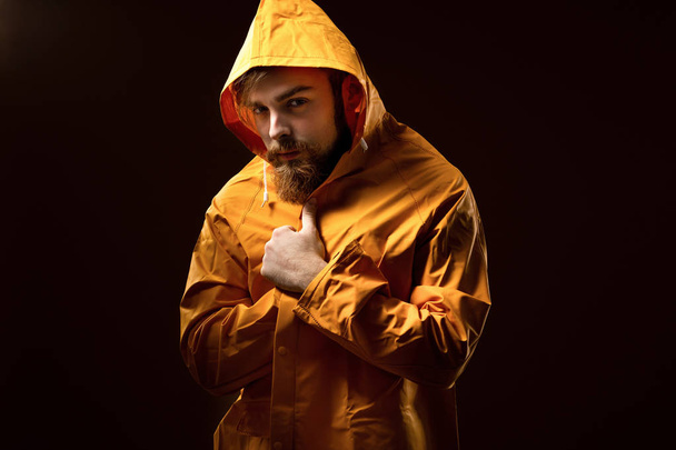 Guy with a beard and mustache dressed in a gray t-shirt and yellow jacket with a hood is standing on a brown background in the studio - Fotografie, Obrázek
