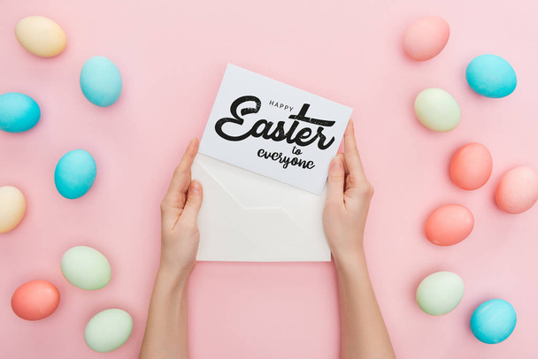bijgesneden beeld van vrouw Holding envelop en wenskaart met Happy Easter aan iedereen belettering in de buurt van veelkleurige geschilderde kip eieren op roze achtergrond  - Foto, afbeelding