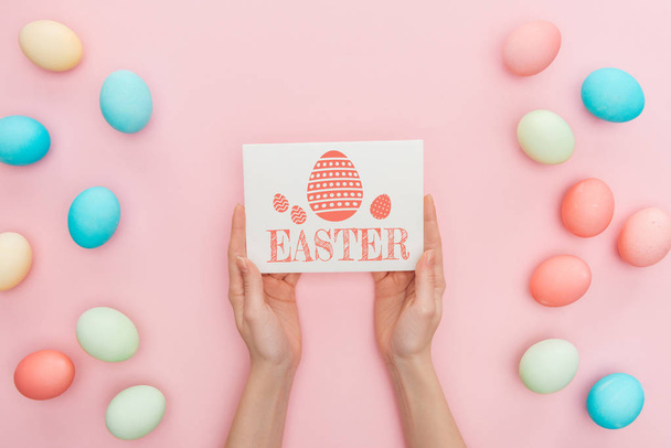 cropped view of woman holding greeting card with Easter lettering near multicolored painted chicken eggs on pink background  - Valokuva, kuva