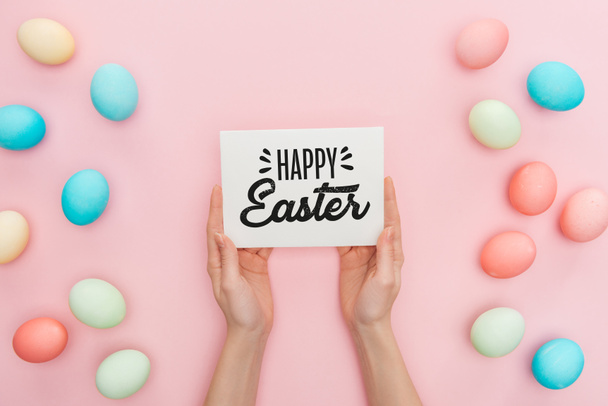 cropped view of woman holding greeting card with happy Easter lettering near multicolored painted chicken eggs on pink background  - Foto, afbeelding