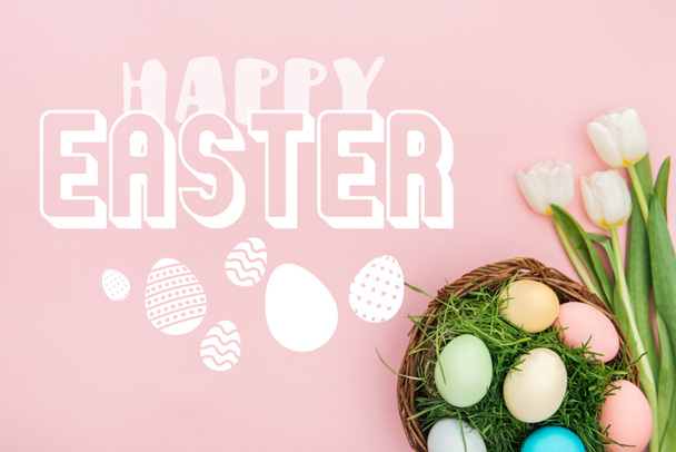 top view of painted chicken eggs on green grass in wicker basket and white tulips on pink background with white happy Easter lettering  - Photo, Image