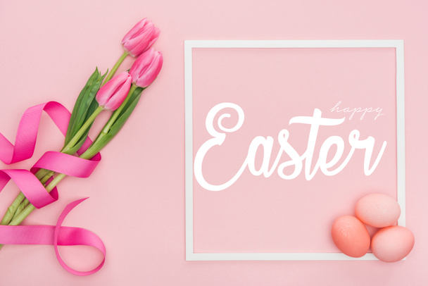 top view of pink tulips bouquet with ribbon and painted eggs near frame with happy Easter lettering on pink background - Photo, Image