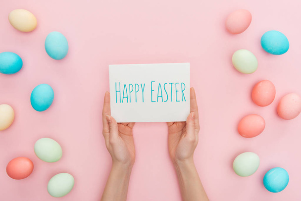 partial view of woman holding greeting card with happy Easter lettering near multicolored painted chicken eggs on pink background  - Foto, Imagen
