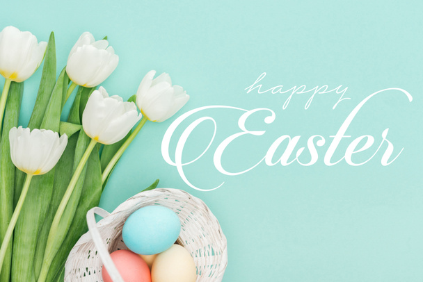 top view of painted chicken eggs in wicker basket and white tulips on blue background with white happy Easter lettering  - Фото, изображение