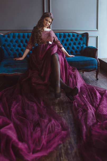 portrait of beautiful young pretty cute woman princess (queen) in long purple queen's dress and crown, with long hair and make up sitting on the blue velvet sofa indoor in minimalistic interior - Foto, immagini